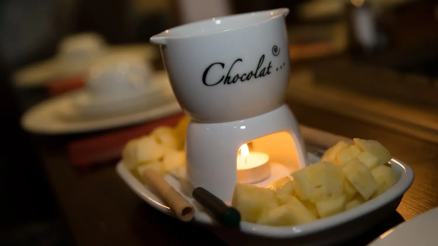 Ein Schokoladenfondue-Set mit einem Keramiktopf mit der Aufschrift „Chocolat..“, der von einer Kerze erhitzt wird, und einer Platte mit geschnittenem Obst (wahrscheinlich Ananas) zum Dippen. Lava Das Indoor BBQ Restaurant